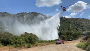 A helicopter drops water near a hillside during a wildfire exercise, as firefighters watch nearby