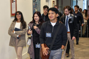 A young man carrying a brown cross-body bag and wearing a dark colored quarter zipper sweater and gray pants smiles as he walks down a hallway. A woman behind him is laughing and a woman beside her is smiling.