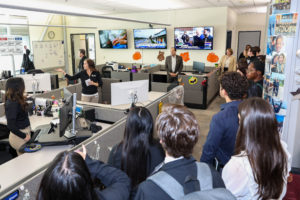 A woman motions to something behind her as a group of people listen to her.