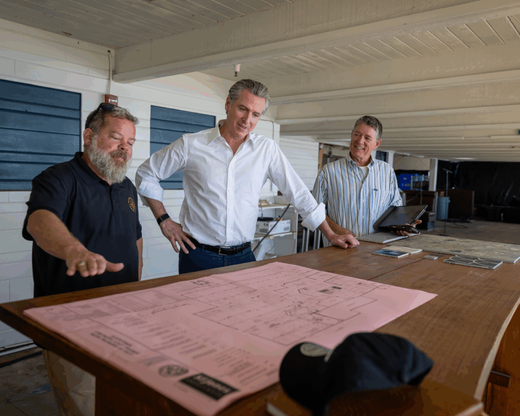 Governor Newsom stands between two men looking at plans.