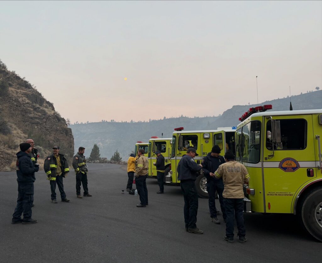 Firefighters stand in front of yellow fire trucks.